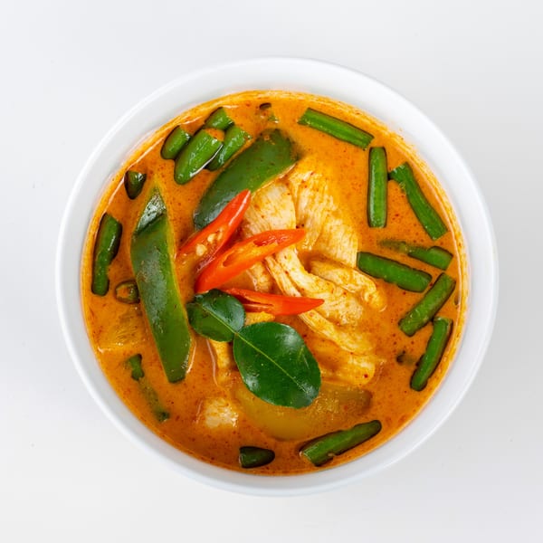 Red curry with chicken, green beans, red chili, and kaffir lime leaves in a white bowl.