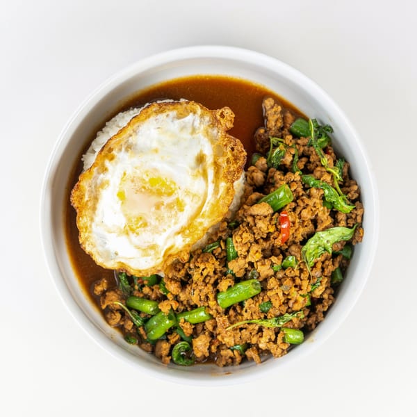 A white bowl of Thai basil chicken with rice, a fried egg, and a rich brown sauce.