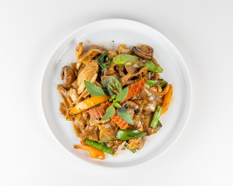 Pad See Ew with chicken, wide noodles, mushrooms, bell peppers, and basil on a white plate.