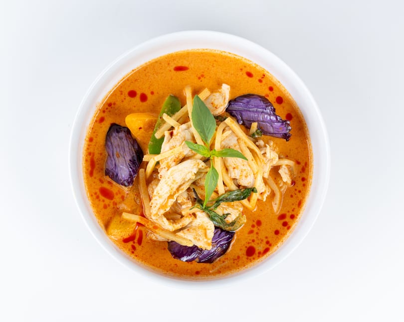 Red curry with chicken, bamboo shoots, eggplant, and basil in a white bowl.