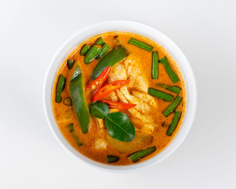 Red curry with chicken, green beans, red chili slices, and kaffir lime leaves in a white bowl.
