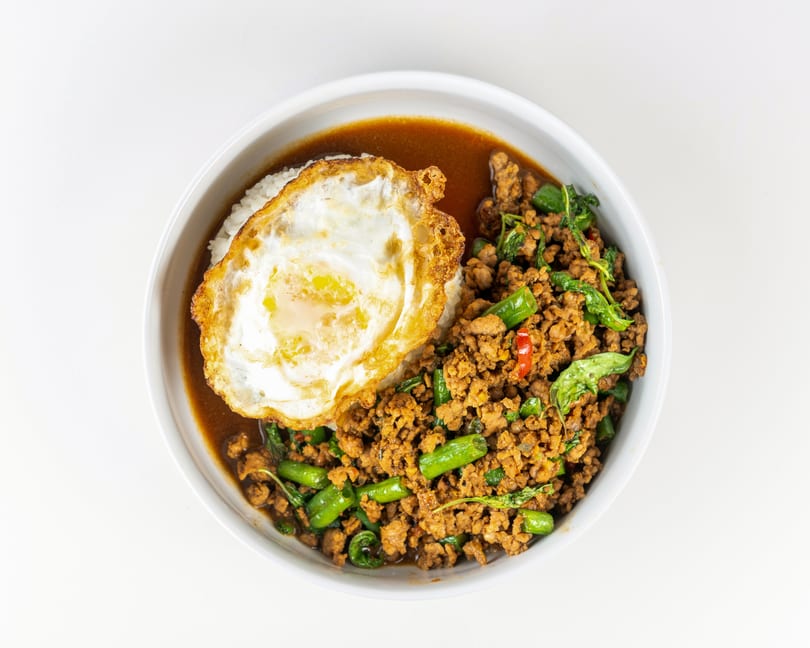 A white bowl of Thai basil chicken with rice and a fried egg on a white background.