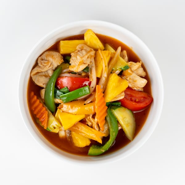 Sweet and sour chicken with pineapple, bell peppers, onions, carrots, and tomatoes in a white bowl.