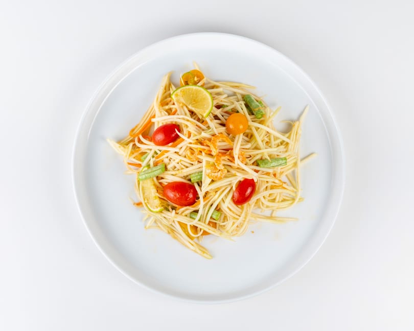 Som Tum, a Thai green papaya salad with tomatoes, green beans, and dried shrimp on a white plate.