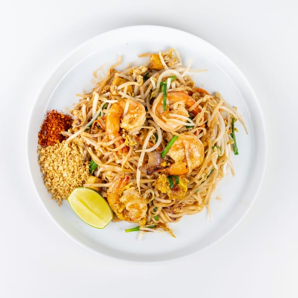 Pad Thai with shrimp, bean sprouts, peanuts, chili flakes, and a lime wedge on a white plate.