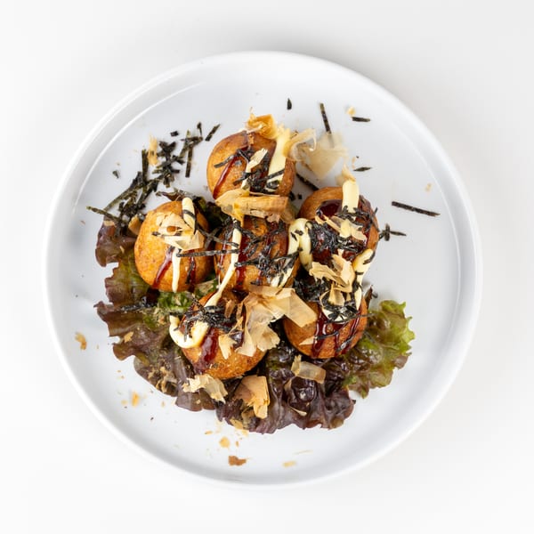 Six takoyaki balls on lettuce, drizzled with sauce, mayonnaise, seaweed, and bonito flakes.
