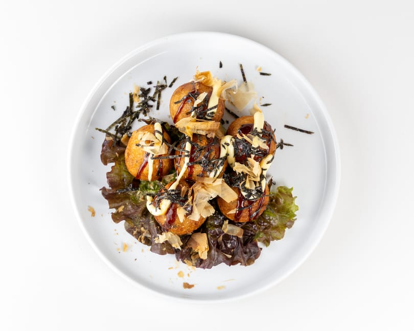 Six takoyaki balls on lettuce, drizzled with sauce, mayonnaise, seaweed, and bonito flakes.